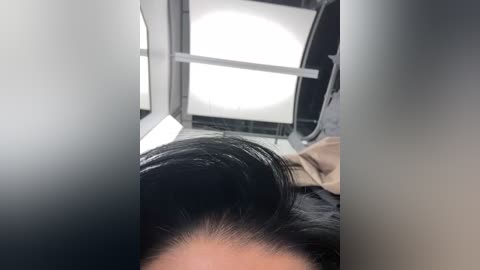 Media: Video of a woman's head, focusing on her black hair styled in a bun. Background shows a dimly lit room with a white light source and grey walls.