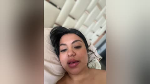 Media: A close-up video of a woman with medium skin tone and black hair, lying on a bed with white pillows and a beige headboard. She has full lips slightly parted, eyes closed, and a relaxed expression.