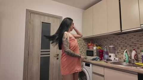 Media: Video of a young woman with long black hair, wearing a sleeveless peach dress, standing in a modern kitchen, washing her hands at a sink, surrounded by appliances and a tiled backsplash.