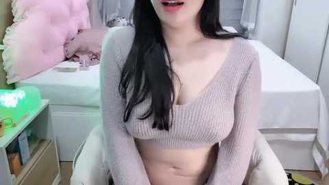 Media: Video of an Asian woman with long black hair, wearing a light pink knit crop top, sitting on a white chair in a cozy, tidy bedroom with a bed, pink and white pillows, and a nightstand.