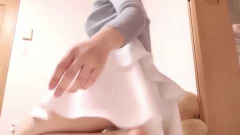 Media: Video of a woman in a light gray sweater and white skirt, captured from a low angle, showing her bent over and lifting her skirt. The background features a beige door and wall.