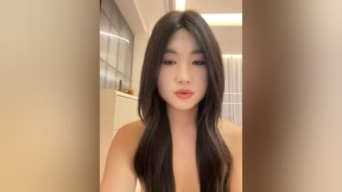 Media: Video of an East Asian woman with long, straight, dark hair, fair skin, and full lips. She is topless, wearing no makeup, in a modern, well-lit room with beige walls and a white window.