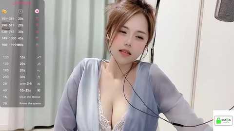 Media: Video of an Asian woman with light skin, brown hair in a messy bun, wearing a light blue cardigan and a lacy bra, sitting with headphones on, in a room with a green curtain and digital time display.