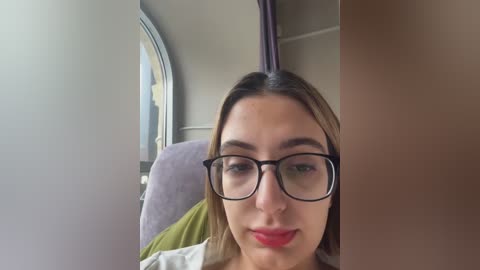 Media: Video of a woman with light skin, brown hair, and black-framed glasses, seated in a train, wearing a green shirt, looking forward.