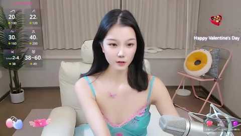 Media: Video of an East Asian woman with long black hair, fair skin, wearing a blue lace bra, sitting in a modern room with a white chair, pink fan, and Valentine's Day-themed decorations.