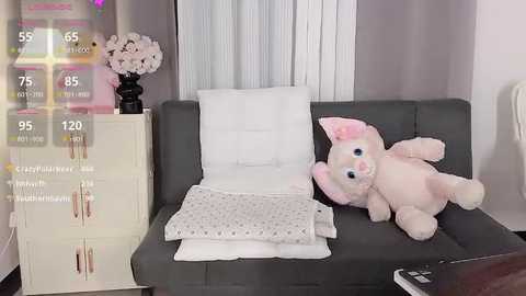 Media: A video of a cozy, modern living room featuring a grey sofa, plush pink bunny, white throw pillows, and a white nightstand with drawers.