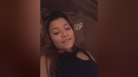 Media: Video of a smiling, fair-skinned woman with long brown hair, wearing a black sleeveless top, lying on a bed with patterned pillows, dimly lit room in the background.