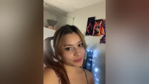 Media: A video of a young woman with light brown hair and light skin, wearing a black spaghetti-strap top, sitting in a dimly lit room with posters on the wall and fairy lights.