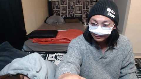 Media: Video of an Asian man with glasses and a black beanie, wearing a gray sweater and a white mask, sitting on a bed with a patterned blanket, in a modest bedroom.