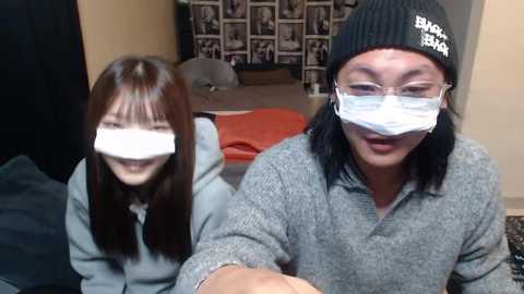 Media: Video of two smiling Asian women, one with straight brown hair, the other with shoulder-length black hair, wearing gray sweaters and black beanie hats with clear face masks. Background shows a bed with orange blanket, gray pillow, and a framed photo wall.