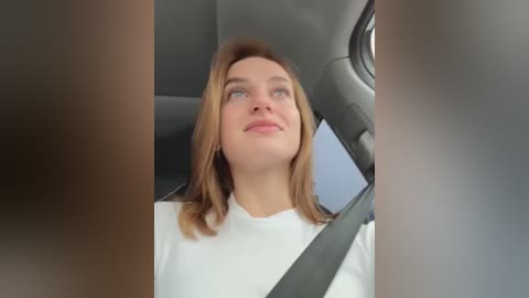 Media: Video of a young woman with light skin and shoulder-length brown hair, wearing a white shirt, seated in a car with a gray seatbelt. She gazes thoughtfully out the window, surrounded by blurred vehicle interiors.