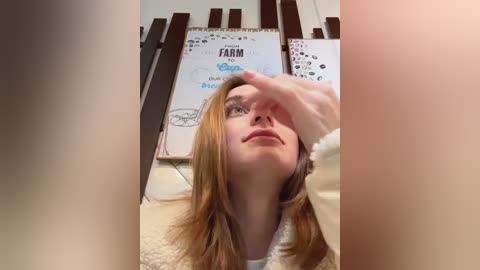 Media: Video of a young woman with light skin, long red hair, and glasses, wearing a white sweater, looking up with one hand over her eyes. Background includes a whiteboard with text and a wooden fence.