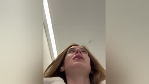 Media: Video of a woman with light skin and straight, shoulder-length blonde hair, eyes closed, mouth slightly open, looking upwards. Background features a beige ceiling and a thin, vertical light strip.