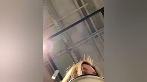 Media: Video of a low-angle view of a blonde woman's head, looking upwards at a high ceiling with exposed beams and industrial lighting, creating a dramatic, slightly distorted perspective.