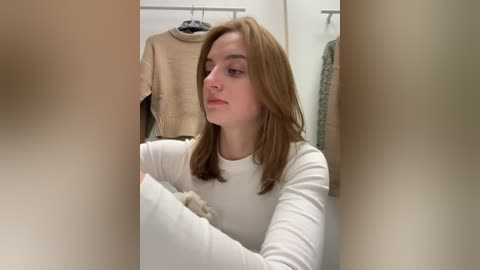 Media: Video of a young woman with long, straight brown hair, wearing a white long-sleeve top, sitting on a beige chair in a minimalist room with hanging clothes on a rack.