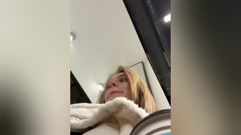 Media: Video of a blonde woman with fair skin, wearing a white, fluffy robe, standing in a modern room with a high ceiling and recessed lights.