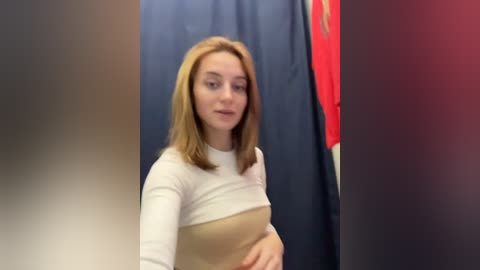 Media: Video of a young woman with straight, shoulder-length blonde hair, wearing a white long-sleeve top and beige vest, standing in a store with navy and red clothing in the background.