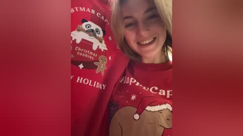 Media: Video of a smiling woman wearing a red Christmas-themed sweater with a cartoon cat and a teddy bear, set against a blurred red background.