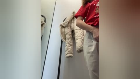 Media: A video captures a person in a red t-shirt and beige pants, holding a beige jacket, standing in a narrow, white-walled hallway with a mirror on the left.