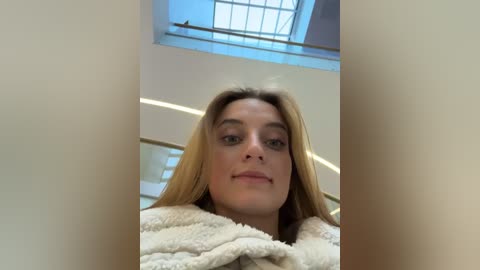 Media: Video of a young Caucasian woman with fair skin and long, straight blonde hair, wearing a fluffy beige robe, standing indoors near a staircase with a glass window.