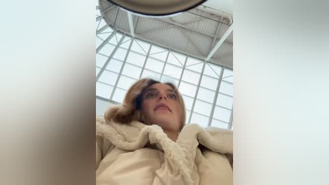 Media: Video of a young woman with light skin, brown hair, and a beige fluffy robe, looking upward in a modern, sunlit room with a high ceiling and large windows.