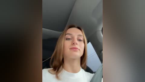 Media: Video of a young Caucasian woman with light skin and straight, shoulder-length brown hair, wearing a white top, seated in a car with grey seats and a white document in the background.