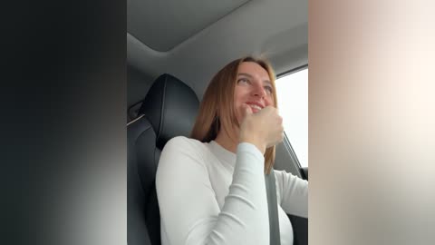 Media: Video of a smiling Caucasian woman with light skin, long brown hair, and a white sweater, sitting in a modern car with black leather seats.