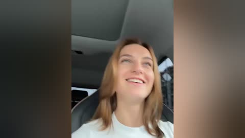 Media: Video of a smiling young woman with straight, light brown hair, wearing a white shirt, seated in a car, with a blurred background showing the car interior.