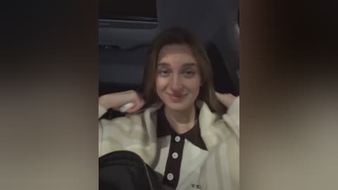 Media: A video shows a young woman with straight brown hair, wearing a beige cardigan over a black collared shirt, smiling and seated in a car.