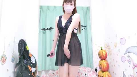 Media: Video of an East Asian woman with light skin, long brown hair, and a slender physique, wearing a sheer black lingerie dress, face mask, and a bat costume, in a Halloween-themed room with decorations.