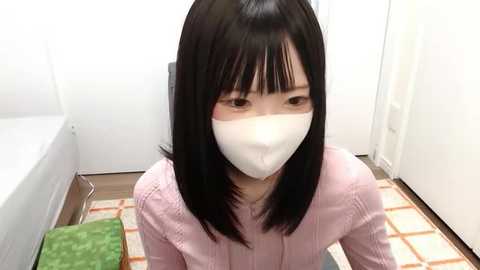 Media: Video of an East Asian woman with straight black hair, wearing a pink sweater and a white face mask, kneeling on a patterned rug in a minimalistic, white-walled room with a bed in the background.