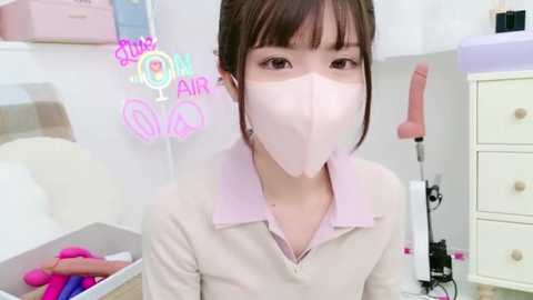 Media: A video of an Asian woman with light skin, wearing a pink face mask and beige sweater, sitting on a bed in a room with a pink vibrator and pastel decor.