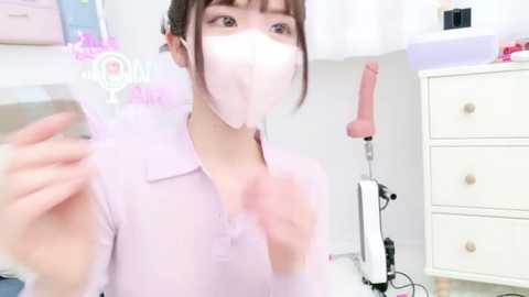 Media: Video of a young East Asian woman in a pastel pink blouse and face mask, standing in a bright, modern room with white furniture and a pink dildo on a shelf.