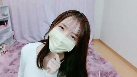 Media: Video of an East Asian woman with long dark hair, wearing a white top and a yellow surgical mask, sitting on a plush pink rug in a minimalist room with white walls and a lavender curtain.