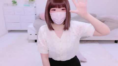 Media: A video of an East Asian woman with straight, shoulder-length brown hair and a white mask, waving in a white room with a bed and white dresser.