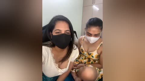 Media: Video of two women, one with dark hair, wearing a white top, black mask, and blue skirt; the other with dark hair, white mask, and floral dress, holding a baby. Background shows tiled wall and doorway.