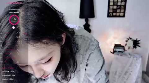 Media: A video of a young Asian woman with long, black hair, wearing a grey cardigan, sitting on a bed with a white sheet. The room is softly lit, featuring a lamp, a nightstand with a clock, and a framed photo on the wall.