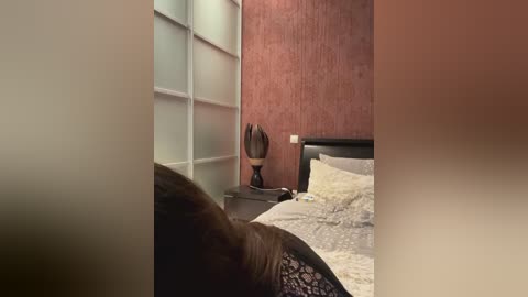 Media: Video of a modern bedroom with beige walls, a dark headboard, and a textured red wall. A bed with white sheets and a decorative pillow is visible, along with a black nightstand holding a decorative lamp.