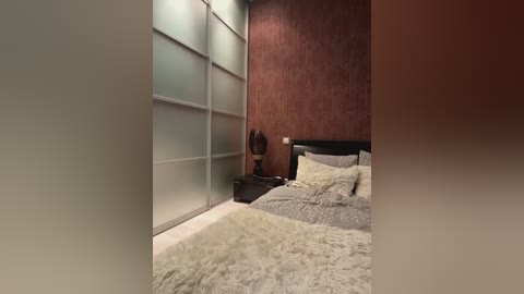 Media: Video of a modern, dimly lit bedroom with frosted glass sliding closet doors, a textured brown wall, a plush white bedspread, and a dark wooden nightstand with a decorative lamp.