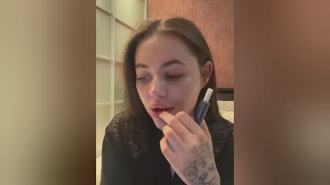 Media: A video of a young woman with light skin, straight dark hair, and a black lace top, applying lipstick. She has a tattoo on her left hand. Background shows a tiled bathroom wall and a wooden door.