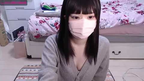 Media: A video shows an Asian woman with long black hair, wearing a pink surgical mask, kneeling on a patterned rug in a bedroom with a floral bedspread and white dresser.