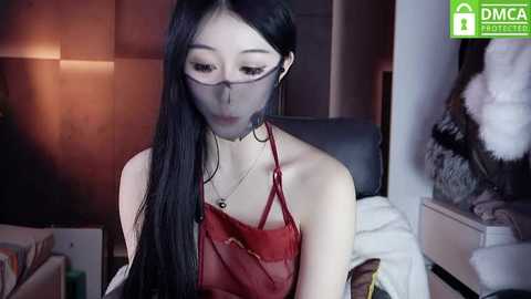 Media: Video of an Asian woman with long black hair, wearing red lingerie and glasses, seated in a dimly-lit room with a bed and white fur coat in the background.