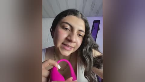 Media: Video of a Latina woman with long dark hair, holding a bright pink vibrator, smiling in a casual white tank top, indoors with a wooden ceiling and purple wall.