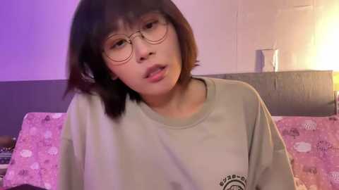 Media: A video of a young Asian woman with short, dark hair and glasses, wearing a grey T-shirt, sitting on a bed with pink floral sheets. The background features a dimly lit room with a wooden headboard and a lamp.