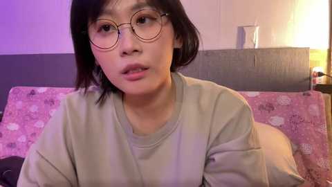 Media: Video of a young Asian woman with short black hair and glasses, wearing a light gray T-shirt, sitting on a bed with pink-patterned pillows. Background includes a headboard and a bedside table with a lamp.