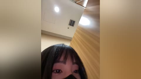 Media: A low-angle, selfie-style video of a woman with dark hair, bangs, and a black mask, captured in a dimly lit room with beige walls and a ceiling fan.