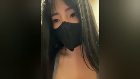 Media: Video of an Asian woman with long black hair and fair skin, wearing a black face mask, covering her mouth and nose, with a blurred, dark background.