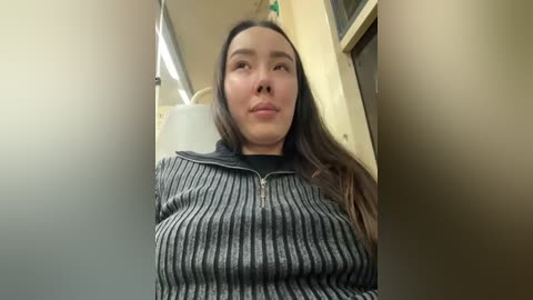 Media: A video of a young Asian woman with long, straight black hair, wearing a black and white striped sweater, seated in a dimly lit subway car, looking slightly upward.