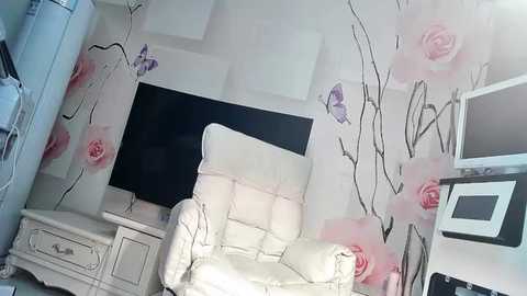 Media: A video of a minimalist, white leather recliner chair in a small, modern room with a white wall adorned with large, pink rose and black vine decals. A flat-screen TV is mounted on the wall, and a white side table with drawers is to the left.