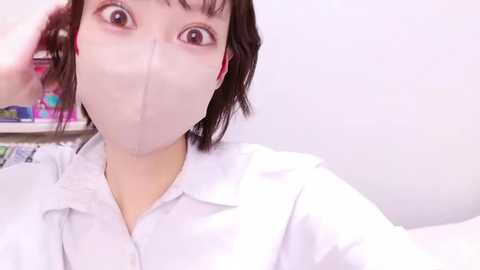 Media: Video of a woman with short dark hair, wearing a white lab coat and face mask, adjusting her hair in a brightly lit room. Background includes shelves with colorful items.